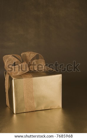 Box wrapped in golden paper with a handmade bow on top.  On a background of shiny gold paper.