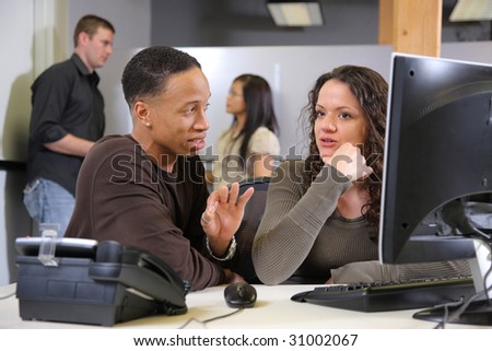Two businesspeople at computer in foreground with co-workers in background