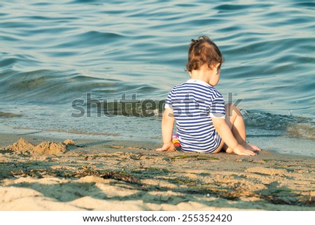 Adorable toddler with sailor shirt sitting at the edge of the waves on a beach. Photo with untraditional color rendering for artistic look