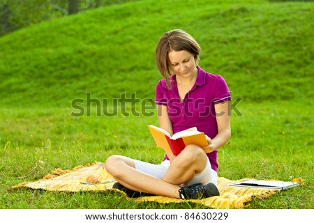 Beautiful woman sitting on the ground in a park and enjoying a funny book. Horizontal shot with copy space.