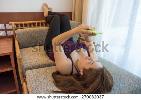 Cheerful Thai woman is texting and chatting with her mobile phone on a sofa in a room