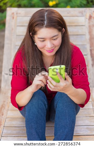 Happy asian woman is playing and chatting on her mobile phone to communication with another people in sunbed wooden seat