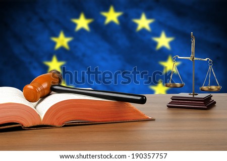 Still life photo of a gavel, scales of justice and law book on a judges bench with the European Union flag behind.