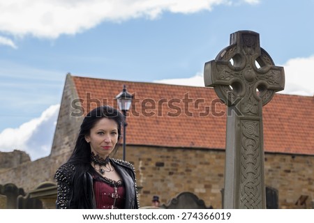 WHITBY, ENGLAND - April 25, 2015: Portrait of a Gothic Girl with a Celtic Stone Cross in a Cemetery. Whitby Goth Weekend is a Twice-Yearly Music Festival for Goths, in Whitby, North Yorkshire, England