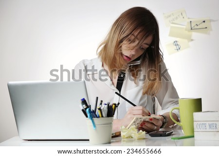 Too busy, Young woman talking on mobile phone and writing notes while sitting at her desk. Pretty caucasian female working in home office.