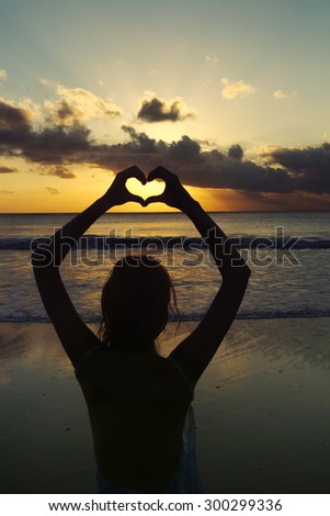 Silhouette of a young woman creating the shape of a heart with her hands against sunset background