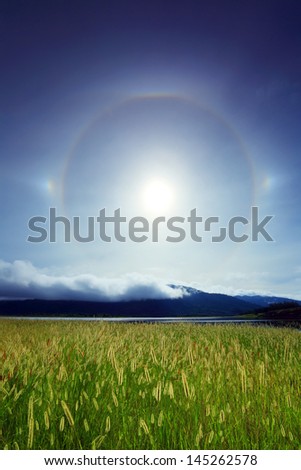 Amazing sun halo above meadow with lake and mountains