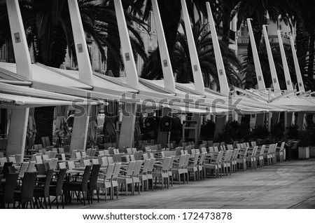 Chairs sit under shade on a promenade in this black and white image. There are no people in this photograph
