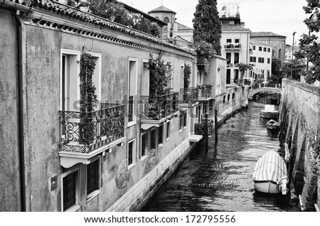 A black and white landscape image of a canal in Venice, Italy with boats moored along the side. A foot bridge is seen and is reflected in the still water