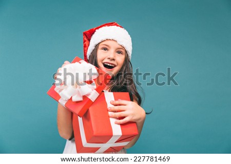 Smiling funny child (kid, girl) in Santa red hat. Holding Christmas gift in hand. Christmas concept. Shooting on blue background