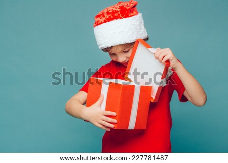 Smiling funny child (kid, boy) in Santa red hat. Holding Christmas gift in hand. Christmas concept. Shooting on blue background