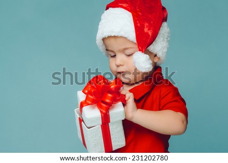 Smiling funny child (kid, boy) in Santa red hat. Holding Christmas gift in hand. Christmas concept. Shooting on blue background