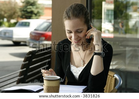 Young woman smiling while doing paperwork and talking on cell phone at cafe.