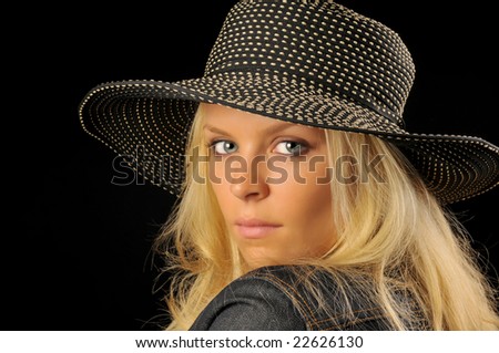 Beautiful blonde woman with spotted hat and denim jacket looking over shoulder with black background.