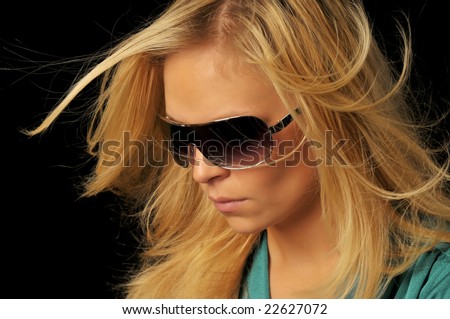 Portrait of beautiful blonde woman with dark sunglasses looking downward left with hair blowing in the wind on black background.