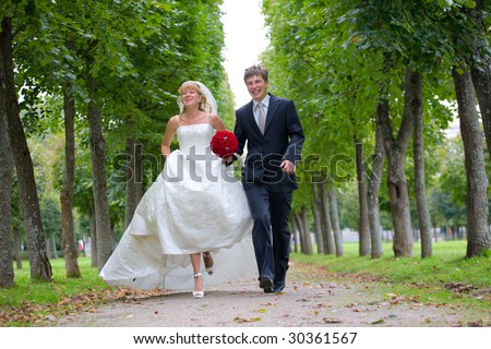Just married couple is walking fast down the path in the park. They are laughing and the bride is raising a little her gown