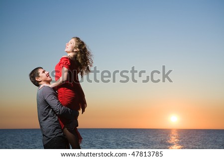 Couple of young adults in love by the sea at sunset. Man holding a woman in his arms.