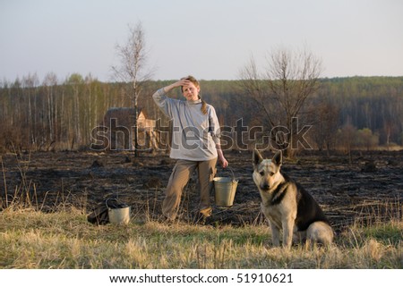 Mature woman with dog and bucket standing at sunset at burned field after extinguish fire. Country house at background.