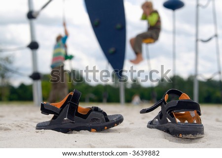 A pair of children-outdoor-shoes standing in the sand of a playground with climbing kids.