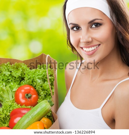 Portrait of happy smiling young beautiful woman in fitness wear holding grocery shopping bag with healthy vegetarian food, outdoors