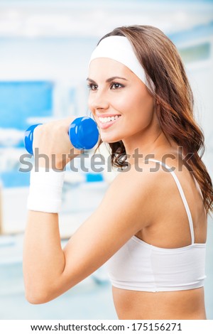 Cheerful woman in fitness wear exercising with dumbbell, at fitness center or gym