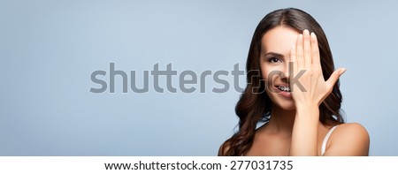 Concept photo of happy smiling young woman with one eye, closed by hand, covering part of her face, over grey, with blank copyspace area for text or slogan