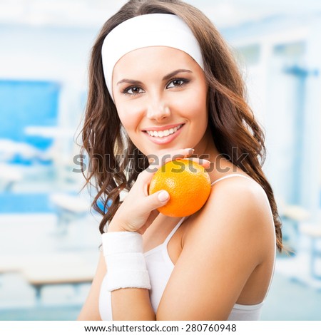 Portrait of smiling young beautiful woman with orange, at fitness center or gym