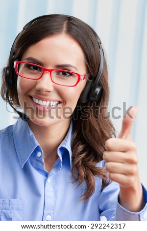 Portrait of happy smiling cheerful young female support phone operator in headset, showing thumbs up hand sign gesture, at office. Customer assistance service concept.