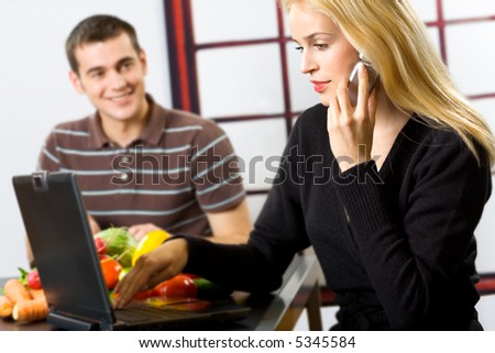 Young happy smiling attractive businesswoman with cellphone and laptop, cooking man at kitchen. Focus on woman.