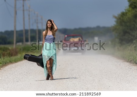 Female walking down a dirt road hitchhiking with a guitar case