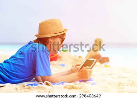 father looking at touch pad while son play with sand at beach