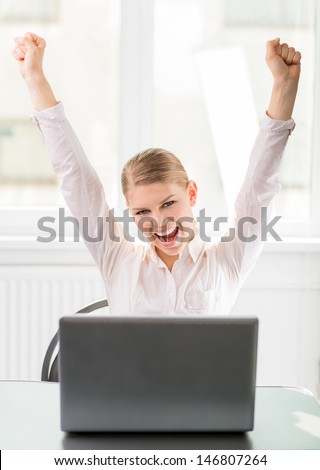 Winning success business woman with hands up at work. Happy female economist with laptop on the desk being glad for company's profit.