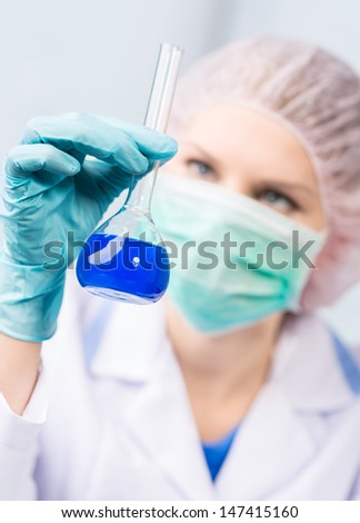 Portrait of science woman looking at blue chemical liquid in lab. Young female chemist in professional uniform making scientific research.