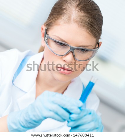 Close-up of female technician woman testing liquid sample in laboratory. Young Caucasian biology female in medical gown, gloves and safety glasses.
