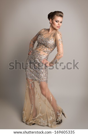 Young blonde in expensive evening dress with diamonds posing in studio. Beautiful fashion model wearing luxury clothes and accessories, with evening hairstyle and professional make up.