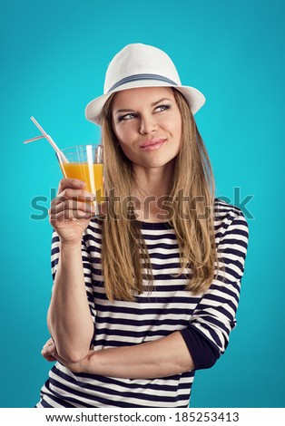 Pretty girl in straw hat standing with juice thinking of summer trip and vacation. Young attractive blond Caucasian female wearing striped clothes posing in studio over blue background.