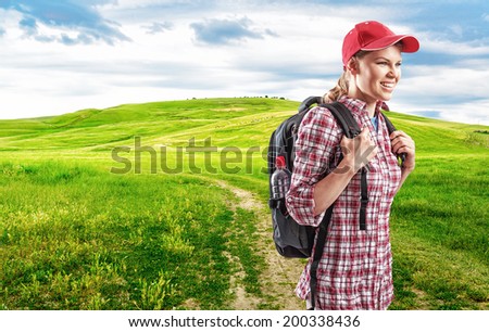 Happy female traveler with backpack walking in countryside. Young woman hiker traveling through green pasture.