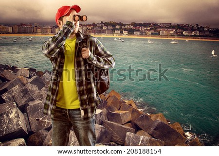 Man climber with backpack looking in binocular at ocean horizon. Young male traveling through sea port.