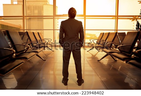 Business man standing in airport hall waiting his flight. Beautiful view of lonely male passenger looking through glass window in airport terminal.