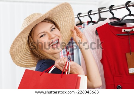 Close-up of beautiful blond lady choosing summer hat standing at clothes rack in shopping mall. Young happy smiling Caucasian woman buying accessories in small boutique.