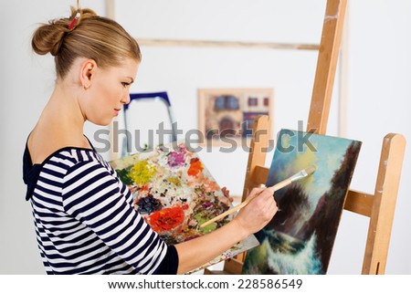 Young female paint artist working with paintbrush over picture. Attractive woman painter standing at easel with color palette.