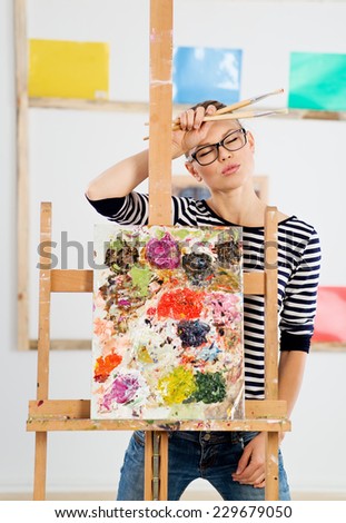 Young woman artist tired after drawing oil picture in her artistic studio. Portrait of professional female painter at work.