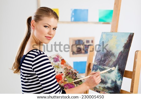 Concentrated student painter standing with color palette and brush at wooden easel. Young artist girl drawing picture in university studio.