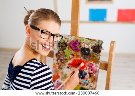 Close-up portrait of attractive cheerful female painter creating new pictorial art. Happy smiling young Caucasian woman with paintbrush standing at easel in her studio.