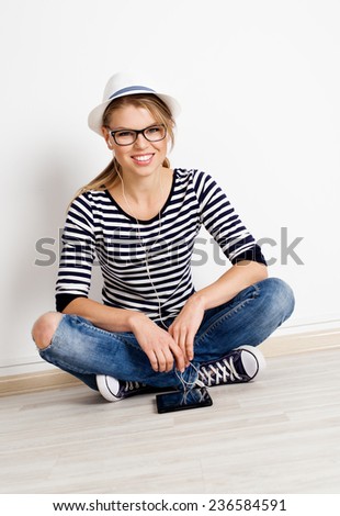 Pretty smiling female sitting at the wall of empty room in headset listening music. Young beautiful woman wearing casual clothes with digital tablet.