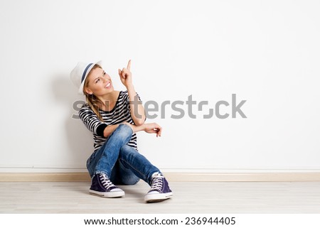 Beautiful woman pointing at copy space on empty wall in new house. Young smiling casual girl sitting on wooden floor and showing smth with her finger.