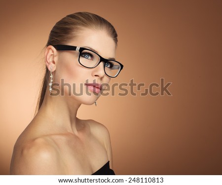 Fashion portrait of young glamour female in optical eyewear with black frame over golden background. Pretty girl with beautiful evening makeup wearing jewelery posing in studio.