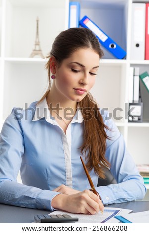 Young female credit risk manager calculating company's risks sitting at her workplace. Young pretty woman accountant with ballpoint.