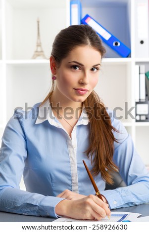 Portrait of young loan consultant signing financial documents in the office. Attractive female professional analyst sitting with ballpoint at the desk.