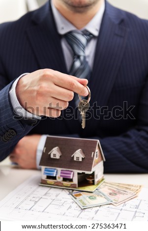 Young businessman broker offering house for rent holding keyring. Closeup of male real estate agent sitting in the office with building model on desk.